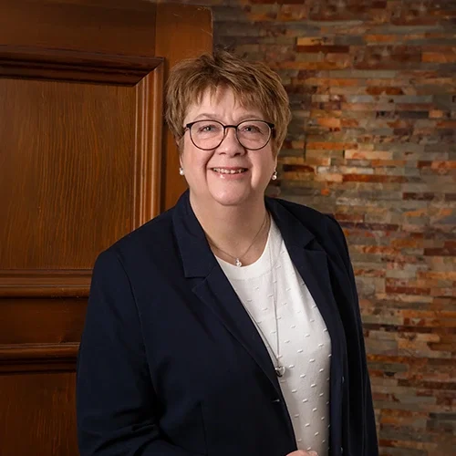 Kvinnlig person med glasögon och kort, brunt hår, klädd i en marinblå blazer och vit topp, står mot en bakgrund av mönstrade stenar. Hon ler och ser ut att vara i en professionell miljö. En träddörr syns delvis bakom henne.