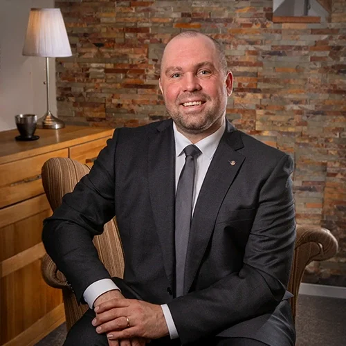 En man i kostym sitter i en fåtölj med en vänlig leende. Han har kort, ljust hår och ett välvårdat skägg. I bakgrunden finns en vägg av sten och ett bord med en lampa och ett glas. Rummet har en varm och inbjudande känsla.