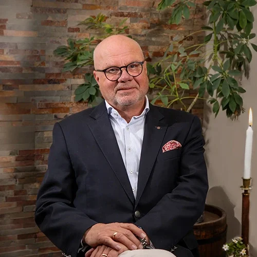 En man med glasögon och skägg sitter i en fåtölj, klädd i en mörk kostymjacka och vit skjorta med en ljusblå skjortkrage. Han har en rosa näsduk i fickan och ser mot kameran med ett vänligt leende. I bakgrunden syns en växt och en vit ljusstake med ett tänd ljus, samt en vägg prydd med stenar i olika nyanser.