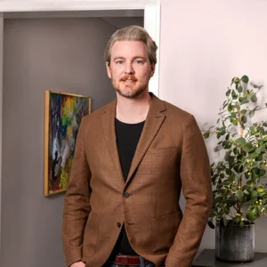 En man i brun kavaj står vid en växt, med en färgstark målning som påminner om den konstnärliga charm som ofta finns i Värnamos begravningsbyråer, vilket förstärker den fridfulla atmosfären.